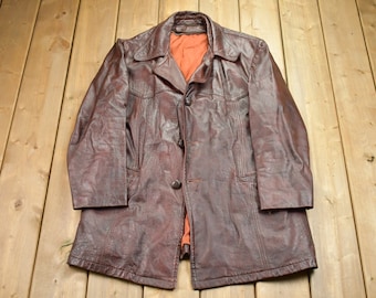 May include: A brown leather jacket with a button-up front and a long, straight cut. The jacket has a slightly textured surface and a dark brown color.