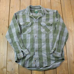 May include: A long-sleeved plaid shirt in green and grey with a button-up front and two chest pockets. The shirt has a label that reads "Cabela's" on the left side of the collar.