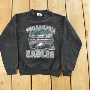 Könnte beinhalten: Schwarzes Sweatshirt mit Rundhalsausschnitt und Philadelphia Eagles Helm-Grafik. Die Grafik enthält den Teamnamen und das Logo in Weiß und Blaugrün. Das Sweatshirt ist aus weichem Material.