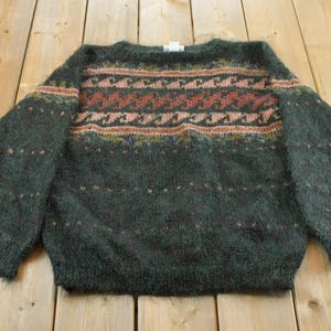 May include: A dark green fuzzy long-sleeve sweater featuring a crew neckline and a horizontal patterned band across the chest. The pattern consists of repeating wave-like shapes in shades of pink, orange, and brown, with small dots throughout.