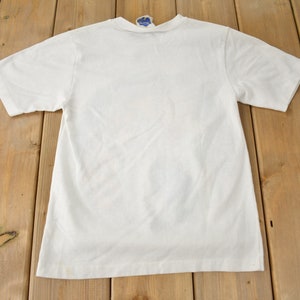 May include: A white t-shirt with a crew neck and short sleeves. The shirt is laying flat on a wooden surface.