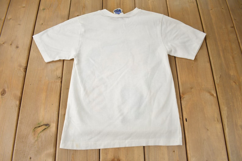 May include: A white t-shirt with a crew neck and short sleeves. The shirt is laying flat on a wooden surface.