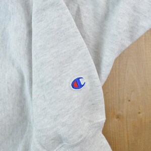 Puede incluir: Una sudadera gris con un logotipo de Champion azul y rojo en la manga izquierda.