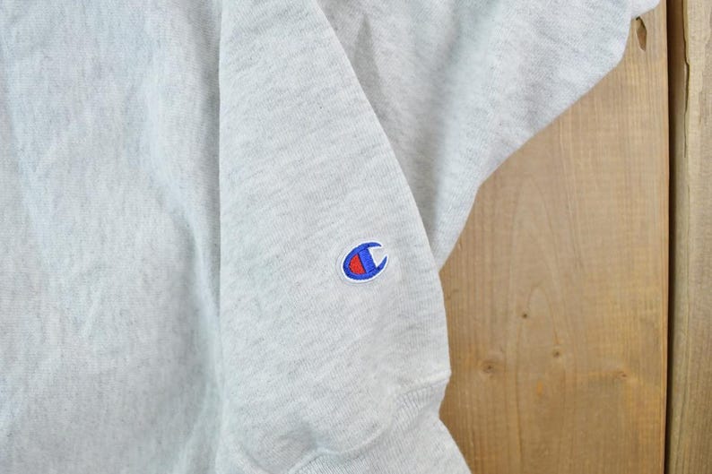 Puede incluir: Una sudadera gris con un logotipo de Champion azul y rojo en la manga izquierda.