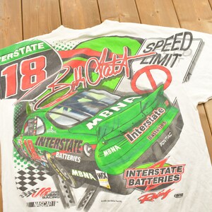 May include: White t-shirt with an all-over print in green and red featuring a racing car, the number 18, and the text "Interstate Batteries" and "MBNA". The shirt also features the signature of "Jeff Gordon" and a speed limit sign.
