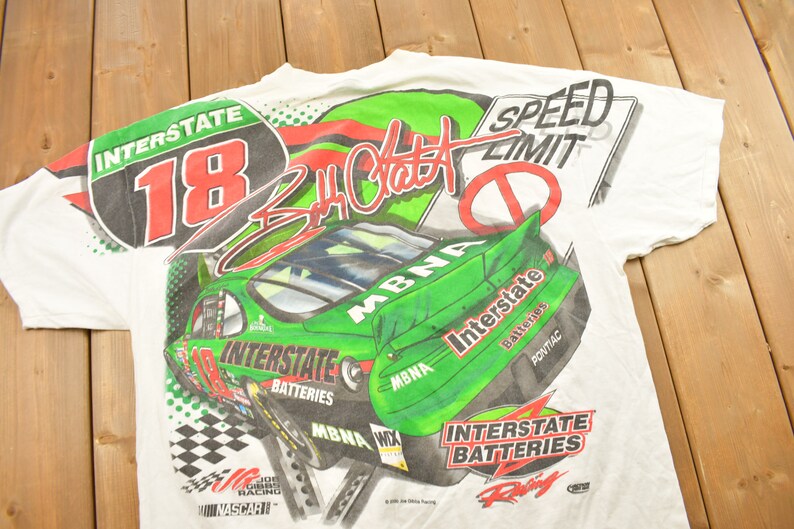 May include: White t-shirt with an all-over print in green and red featuring a racing car, the number 18, and the text "Interstate Batteries" and "MBNA". The shirt also features the signature of "Jeff Gordon" and a speed limit sign.