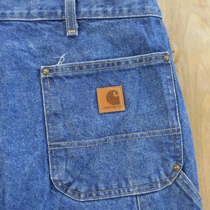 K&ouml;nnte beinhalten: Nahaufnahme einer blauen Jeans-Arbeitshose. Die Hose hat eine gro&szlig;e Tasche mit einem braunen Leder-Carhartt-Patch. Die N&auml;hte sind sichtbar und die Hose scheint abgenutzt zu sein.