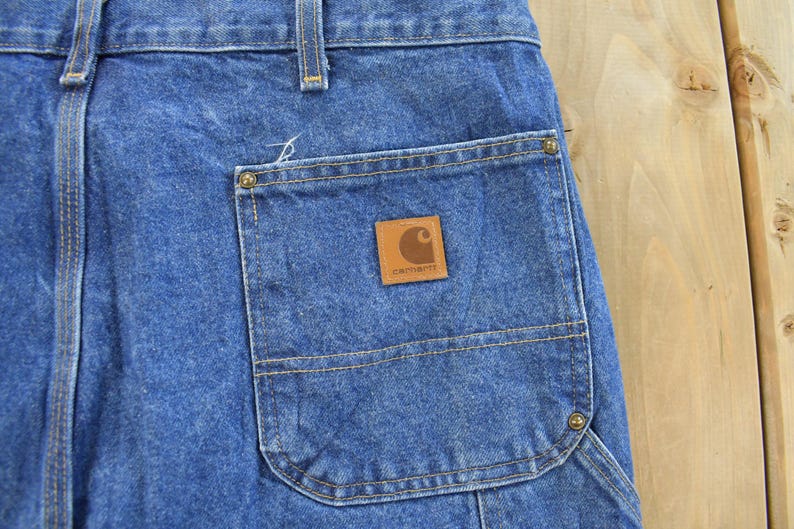 K&ouml;nnte beinhalten: Nahaufnahme einer blauen Jeans-Arbeitshose. Die Hose hat eine gro&szlig;e Tasche mit einem braunen Leder-Carhartt-Patch. Die N&auml;hte sind sichtbar und die Hose scheint abgenutzt zu sein.
