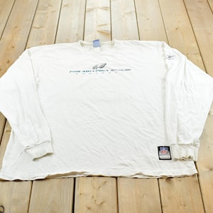 May include: A white long-sleeve t-shirt featuring the Philadelphia Eagles logo and text on the chest. The shirt includes a small Reebok logo on the left sleeve and an NFL patch near the bottom hem. The garment has a crew neckline and ribbed cuffs.