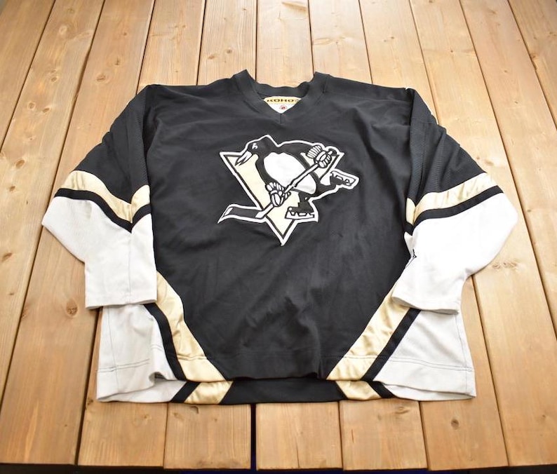 Puede incluir: Camiseta de hockey negra y blanca con detalles dorados. La camiseta presenta el logotipo de los Pittsburgh Penguins con un ping&uuml;ino sosteniendo un palo de hockey. La camiseta tiene mangas largas y cuello en V.