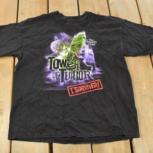May include: Black t-shirt featuring a graphic design for "The Twilight Zone Tower of Terror." The design includes the words "Tower of Terror" in purple and white, a green and white monster, and the text "I Survived!" in a red rectangle.