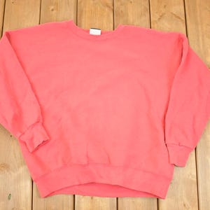 Puede incluir: Una sudadera de cuello redondo de color coral. La parte superior de manga larga tiene puños y dobladillo acanalados. La sudadera está hecha de un material suave y está colocada sobre una superficie de madera. La etiqueta es visible en la parte posterior del cuello.