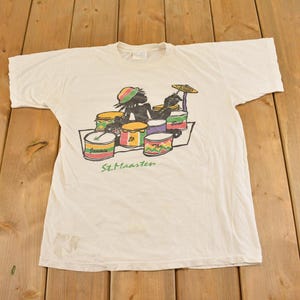 Może przedstawiać: Biały t-shirt z krótkim rękawem z grafiką przedstawiającą czarną sylwetkę grającą na perkusji. Bębny są w kolorach czerwonym, żółtym i zielonym. Pod perkusją znajduje się nadruk z tekstem "Jammin to the Beat" oraz "St. Maarten".