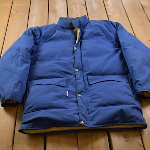 Könnte beinhalten: Eine blaue Daunenjacke mit Druckknopfverschluss und zwei großen Taschen. Die Jacke hat ein braunes Futter.