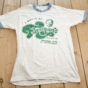May include: White t-shirt with a green and blue logo that says "I got it at  Flanigan's" with a drawing of a man's face. The text below the logo says "Spring of '80 Brother Jim's Ft. Lauderdale."