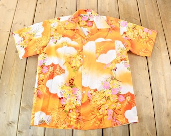 May include: A short-sleeved button-up shirt with a floral print in shades of orange, yellow, and white. The shirt has a pocket on the left side.