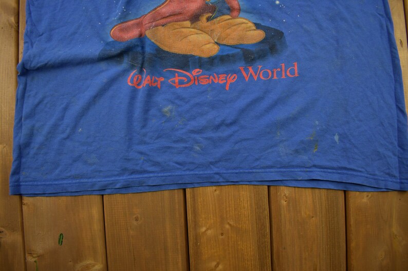 Pu&ograve; includere: Una maglietta blu con un personaggio dei cartoni animati che indossa un cappello rosso e scarpe marroni. Il testo "Walt Disney World" &egrave; stampato sulla maglietta.