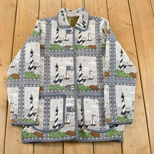 May include: A light-coloured jacket featuring a repeating pattern of lighthouses, sailboats, and coastal scenes. The jacket has a button-up front, a collar, and a blue trim. The overall theme is nautical.