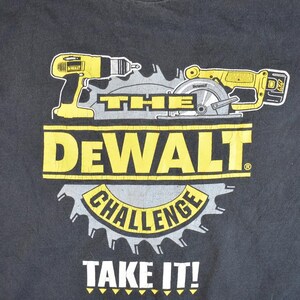 May include: Black t-shirt featuring a yellow and black graphic of a drill and circular saw with the text "The Dewalt Challenge Take It!"