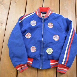 Op de afbeelding: Blauw baseballjack met rode en witte strepen en verschillende baseball-thema patches, waaronder "Big Leaguer", "Come Run", "Twin Athletics Group", "Royals Northwest Shell" en "Home Sweet Home".