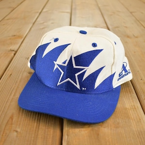 Può includere: Un cappellino da baseball vintage con corona bianca e visiera blu. Il cappellino presenta un grande design a stella blu con dettagli bianchi e la scritta "LOGO ATHLETIC". Il cappello è realizzato in materiale resistente.