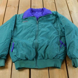 May include: Teal and purple reversible jacket with a zip-up front. The jacket has a purple collar and teal cuffs and hem. The Columbia logo is visible on the chest. The jacket is made of a lightweight material.
