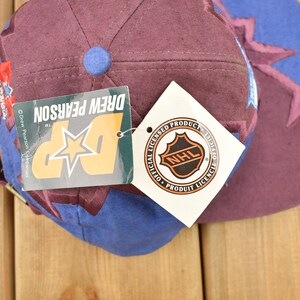 May include: A purple and blue baseball cap with a star and the text "Drew Pearson" on the side. The cap also has a white tag with the NHL logo and the text "Official Licensed Product".