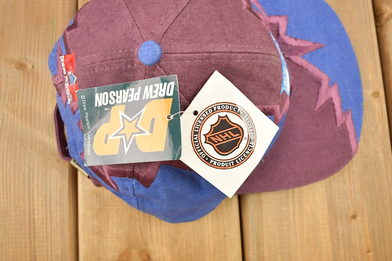 May include: A purple and blue baseball cap with a star and the text "Drew Pearson" on the side. The cap also has a white tag with the NHL logo and the text "Official Licensed Product".