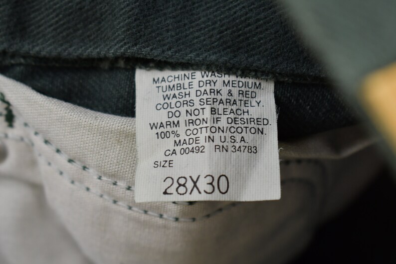 May include: A white care label with black text detailing washing instructions for a pair of jeans. The label reads "MACHINE WASH WARM, TUMBLE DRY MEDIUM, WASH DARK & RED COLORS SEPARATELY, DO NOT BLEACH, WARM IRON IF DESIRED, 100% COTTON/COTON, MADE IN U.S.A., CA 00492 RN 34783, SIZE 28X30".