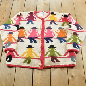 May include: A cream-colored knit cardigan featuring a repeating pattern of stylized figures wearing hats and holding hands. The sweater has a round neckline, long sleeves, a front button closure, and red trim along the edges, cuffs, and hemline.