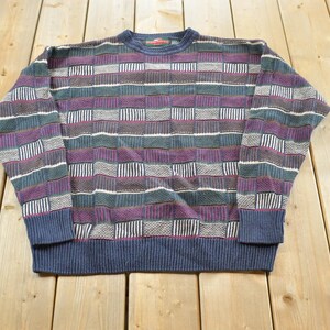 Peut inclure: Un pull vintage à col rond avec un motif géométrique. Le pull présente un motif en damier dans les tons de violet, sarcelle, gris et blanc. Les poignets, le col et l'ourlet sont bleu marine uni. Le pull est en maille douce.