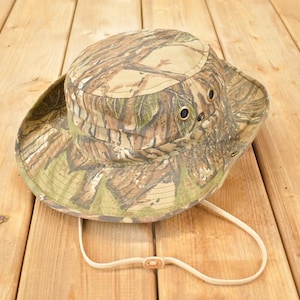 Op de afbeelding: Camouflage vissershoed met een brede rand en een kinband. De hoed heeft een bos camouflagepatroon in groen, bruin en beige tinten. Het heeft oogjes voor ventilatie en een neutrale kinband.