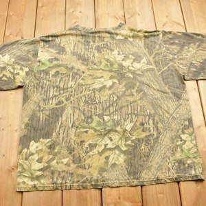 May include: A camouflage pattern t-shirt with a crew neck and short sleeves. The shirt features a woodland camouflage design in shades of brown, green, and tan. The shirt is made of a soft, likely cotton, material.