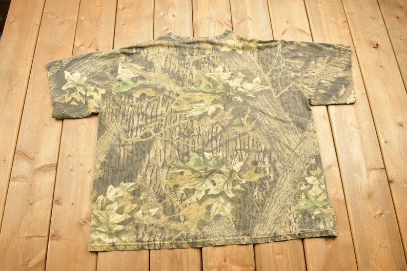 May include: A camouflage pattern t-shirt with a crew neck and short sleeves. The shirt features a woodland camouflage design in shades of brown, green, and tan. The shirt is made of a soft, likely cotton, material.