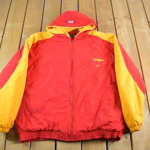 May include: A red and yellow hooded jacket with a full-length zipper. The jacket has yellow sleeves and hood lining, with the word "CHIEFS" and a small Nike logo on the chest. The jacket is made of a lightweight material.