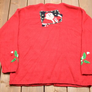 K&ouml;nnte beinhalten: Rote Strickjacke mit einem festlichen Weihnachtsmann-Motiv auf der R&uuml;ckseite und gr&uuml;nen Stechpalmen-Akzenten an den &Auml;rmeln.