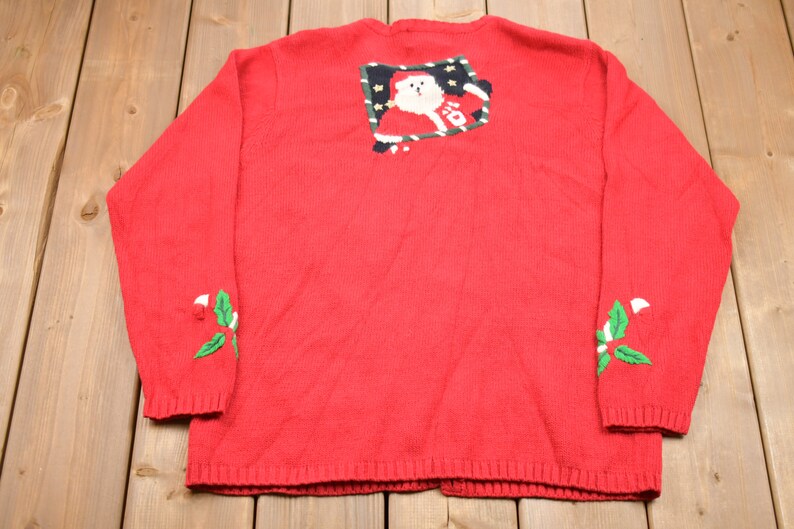 K&ouml;nnte beinhalten: Rote Strickjacke mit einem festlichen Weihnachtsmann-Motiv auf der R&uuml;ckseite und gr&uuml;nen Stechpalmen-Akzenten an den &Auml;rmeln.