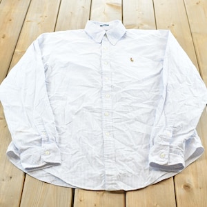 May include: A white shirt with blue vertical stripes, featuring long sleeves and buttoned cuffs. The collar is buttoned. A small logo is embroidered on the chest. The shirt is displayed on a wooden surface.