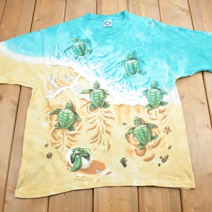 Puede incluir: Una camiseta tie-dye con una escena de playa con tortugas marinas beb&eacute;s. La camiseta es de color azul claro y amarillo con un dise&ntilde;o de tortuga verde y marr&oacute;n. Las tortugas caminan por la arena y nadan en el oc&eacute;ano.