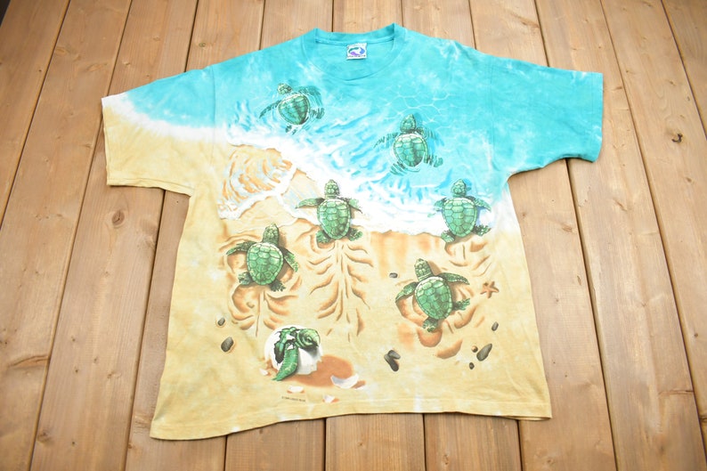 Puede incluir: Una camiseta tie-dye con una escena de playa con tortugas marinas beb&eacute;s. La camiseta es de color azul claro y amarillo con un dise&ntilde;o de tortuga verde y marr&oacute;n. Las tortugas caminan por la arena y nadan en el oc&eacute;ano.