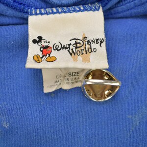 Pu&ograve; includere: Un'etichetta bianca con il testo "Walt Disney World" e un'immagine di cartone animato di Topolino. L'etichetta &egrave; attaccata a una maglietta blu con il testo "One Size" e "Made in U.S.A."