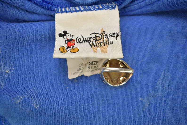 Pu&ograve; includere: Un'etichetta bianca con il testo "Walt Disney World" e un'immagine di cartone animato di Topolino. L'etichetta &egrave; attaccata a una maglietta blu con il testo "One Size" e "Made in U.S.A."