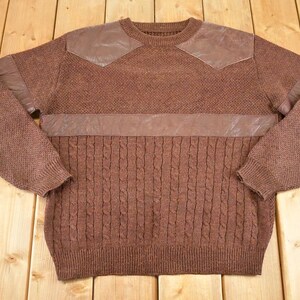Könnte beinhalten: Brauner Pullover mit Lederaufnähern auf den Schultern und einem horizontalen Band über der Brust. Der untere Teil des Pullovers hat ein Zopfmuster, während die Ärmel und die obere Brust ein anderes Strickmuster haben.