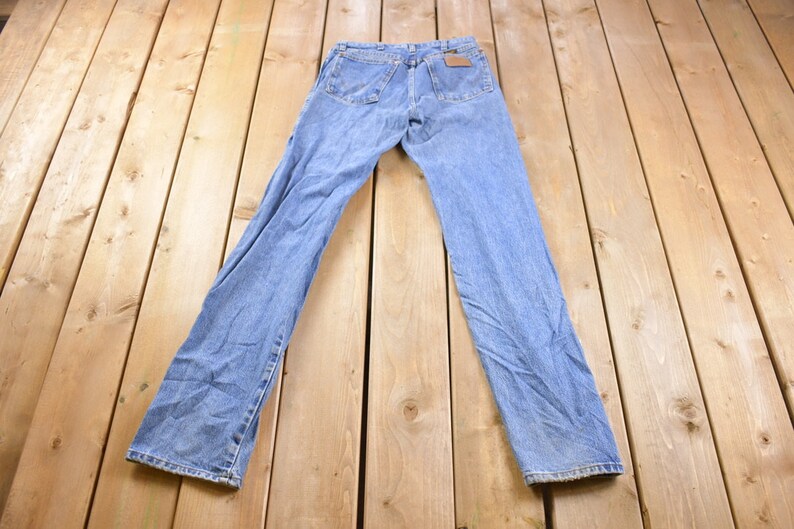 May include: A pair of light blue denim jeans laid flat on a wooden surface. The jeans have a classic straight leg style and a light wash.