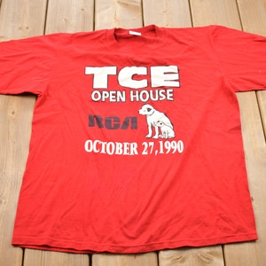 Könnte beinhalten: Rotes T-Shirt mit weißem Text, der "TCE Open House RCA October 27, 1990" lautet. Das Shirt zeigt außerdem eine weiße Illustration von zwei Hunden.