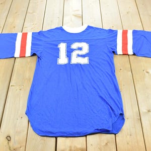 May include: A blue short-sleeved jersey featuring a white crew neckline and the number 12 printed in white on the chest. The sleeves are accented with two horizontal red stripes and one white stripe, creating a classic athletic appearance.