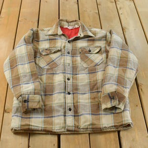 May include: A vintage plaid flannel shirt in shades of brown, tan, and blue. The shirt has two chest pockets, button closures, and a red lining visible at the collar. The shirt is laid flat on a wooden surface.