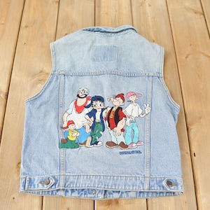 May include: A light blue denim vest with an embroidered patch featuring a group of cartoon characters. The characters are wearing colourful clothing and are standing together. The text "Universal Studios" is visible at the bottom of the patch.