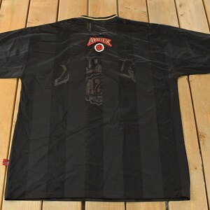 Op de afbeelding: Zwart shirt met verticale strepen. Het shirt heeft rode en witte accenten op de mouwen en kraag. Het Avirex-logo is zichtbaar op de achterkant. Het nummer 42 is zichtbaar op de achterkant van het shirt.