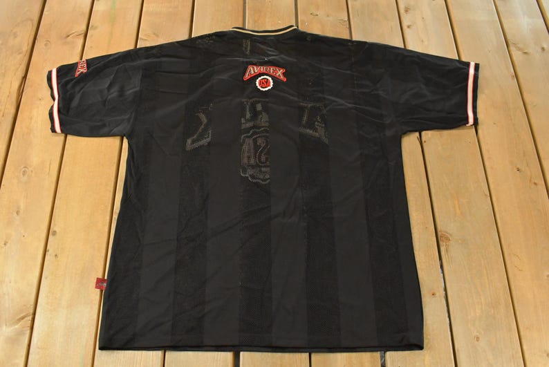 Op de afbeelding: Zwart shirt met verticale strepen. Het shirt heeft rode en witte accenten op de mouwen en kraag. Het Avirex-logo is zichtbaar op de achterkant. Het nummer 42 is zichtbaar op de achterkant van het shirt.
